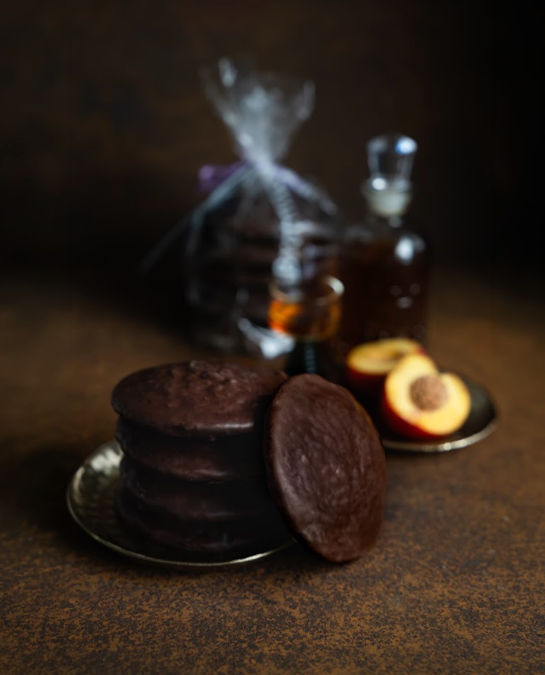 Gestapelte schokolierte Aprikose-Rum-Lebkuchen auf Metalltellerchen, dahinter eine halbierte Aprikose und eine Karaffe Rum.