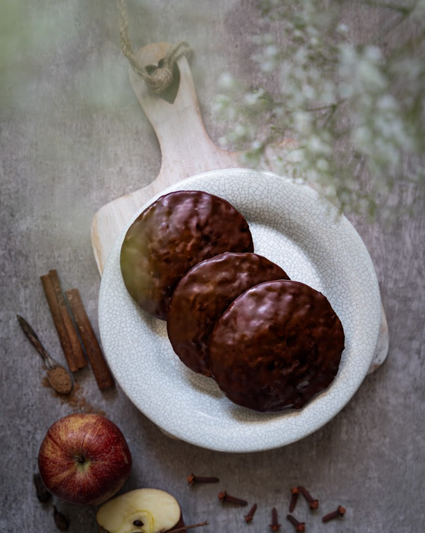 Drei schokolierte Bratapfel-Lebkuchen auf weißem Teller, daneben Zimtstangen, ein Apfel und Nelken