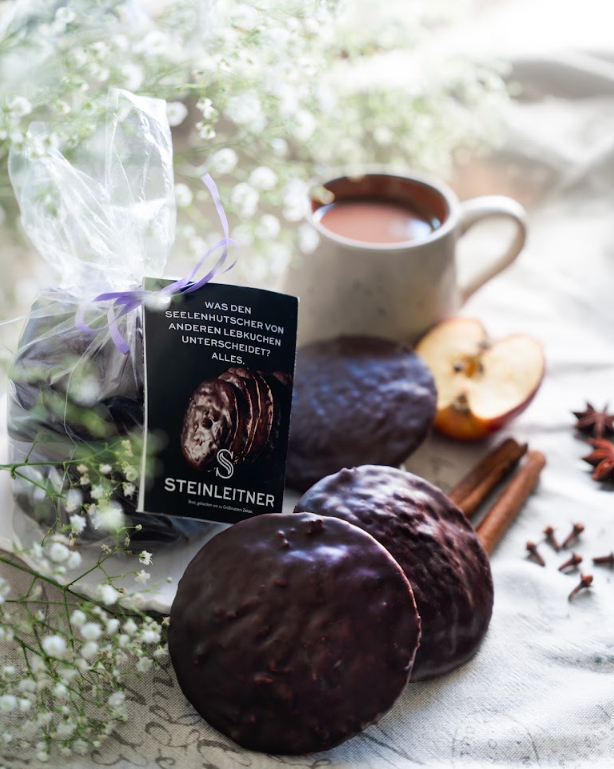 Bratapfel-Lebkuchen von Steinleitner in Geschenktüte mit Anhänger, dahinter eine Tasse Kakao und weihnachtliche Gewürze